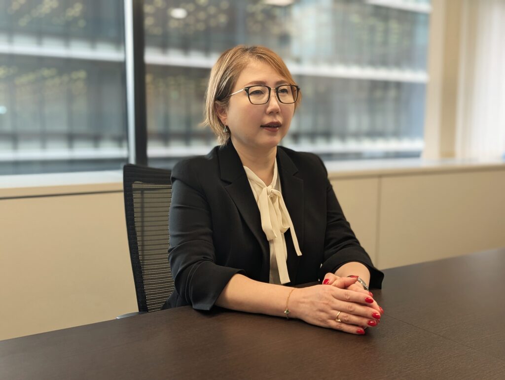 A woman in a black blazer sits at a conference table, hands clasped together, with a professional demeanor and a neutral expression. She is in an office setting with large windows in the background.