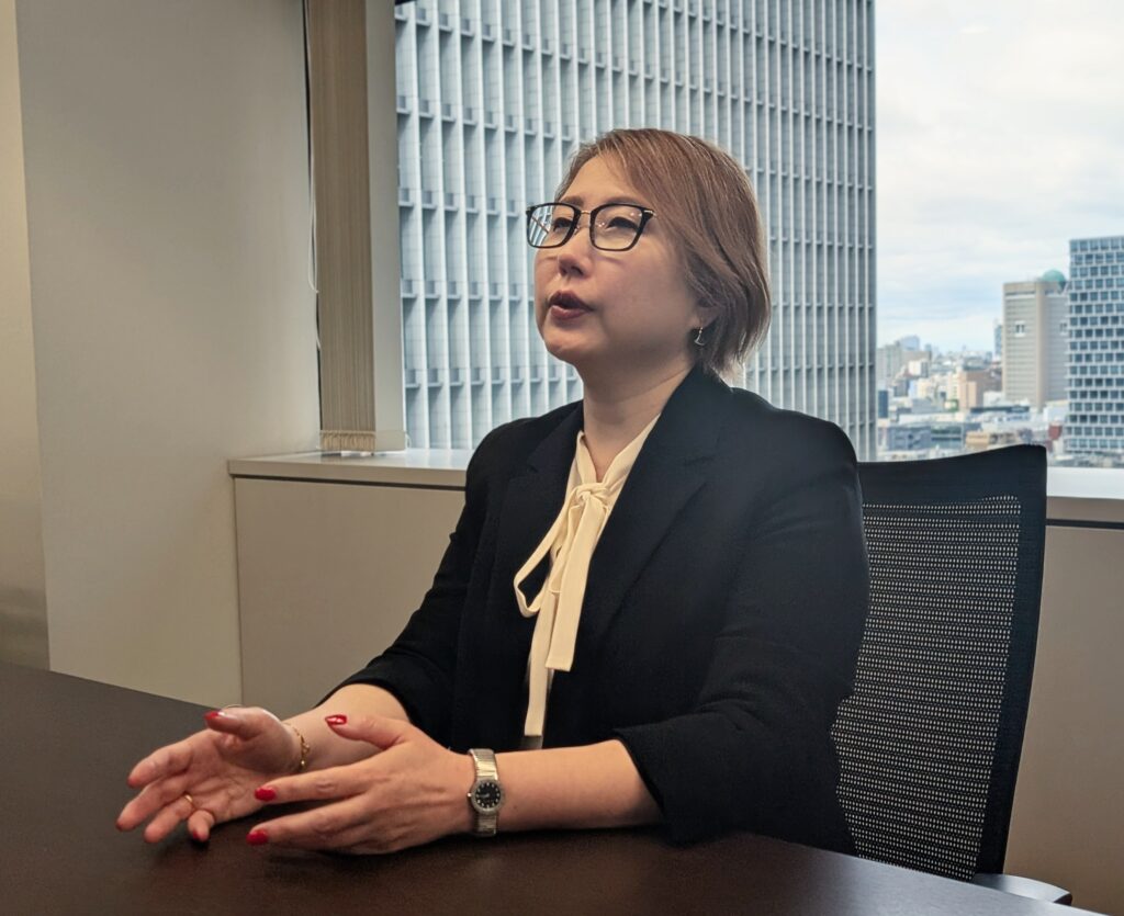 A woman with short brown hair, wearing glasses and a black blazer, sits at a table in a modern office. She appears to be conversing, with her hands gesturing expressively, and a city skyline visible through the window behind her.